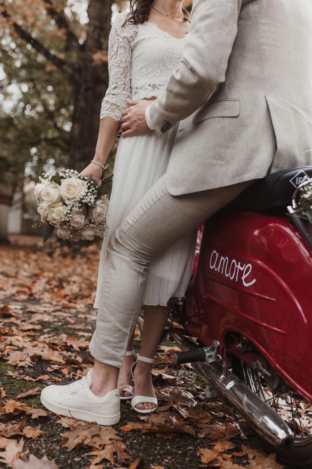Hochzeitspaar umarmt sich in der Nähe eines roten Oldtimer-Motorrads mit weißem Blumenstrauß, während es auf Herbstblättern steht.