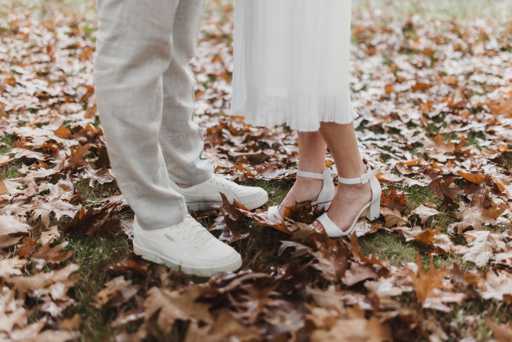 Ein Paar steht in Hochzeitskleidung auf abgefallenem Herbstlaub und zeigt detailliert weiße Anzughosen und glitzernde Schuhe.