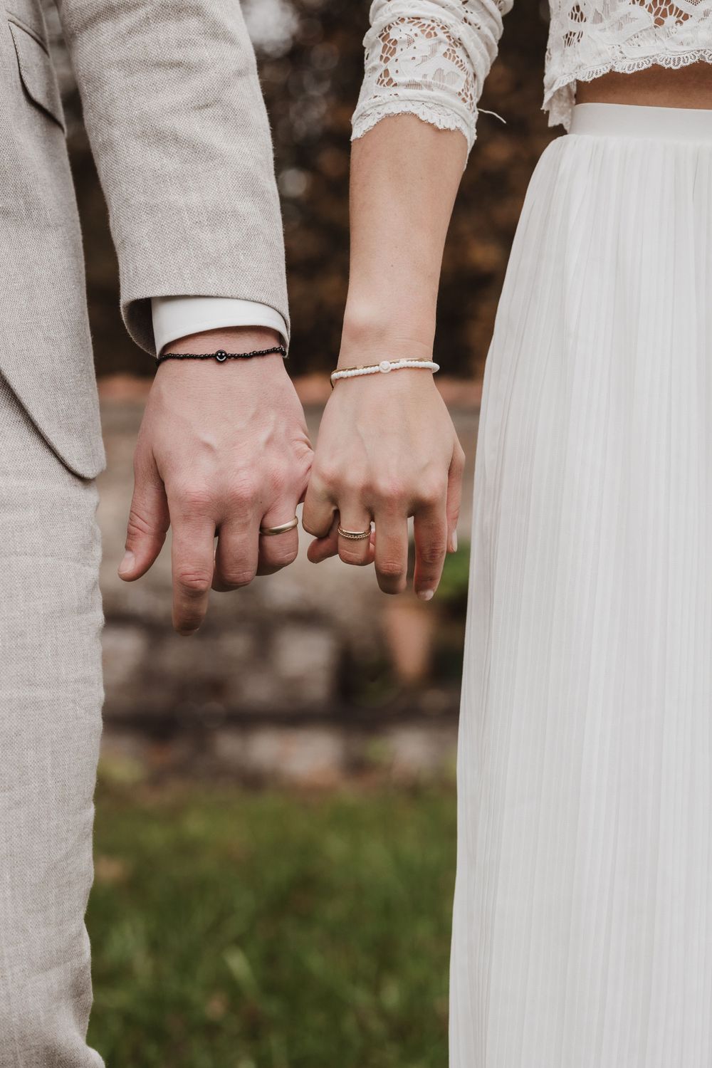 Nahaufnahme eines frisch vermählten Paares, das Händchen hält und Eheringe und weiße Spitzenärmel vor beigem Anzug zeigt.
