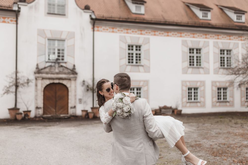 Hochzeitspaar umarmt sich vor einem historischen weißen Gebäude mit verzierten Fenstern und einem großen Eingang.
