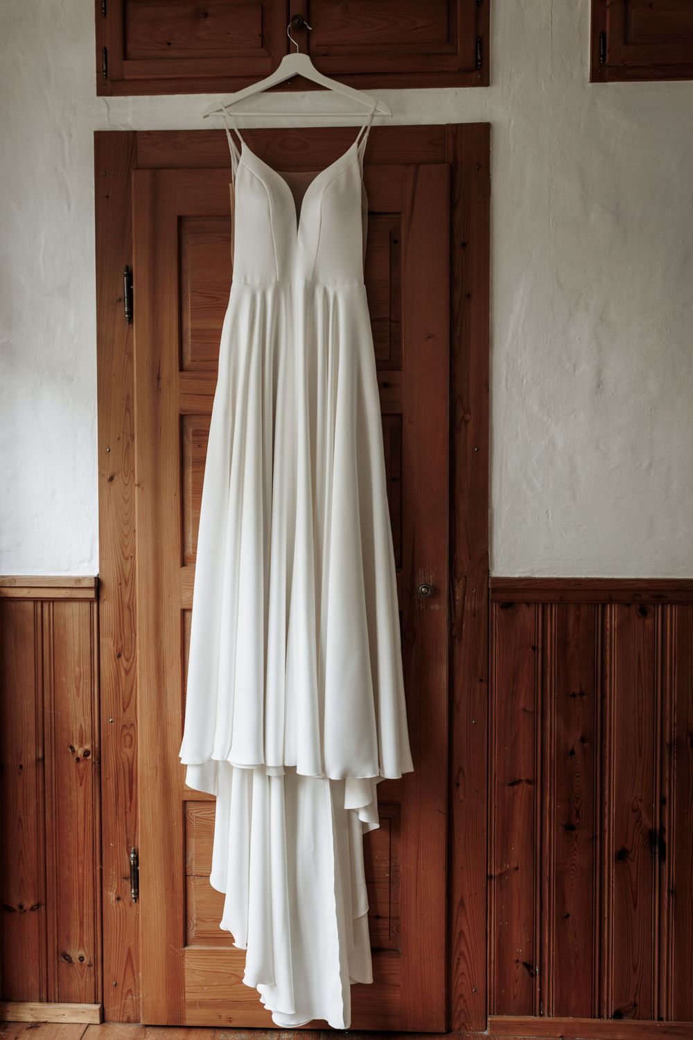 Ein elegantes weißes Brautkleid mit V-Ausschnitt und fließendem Rock hängt an einer Holztür mit rustikaler Täfelung.