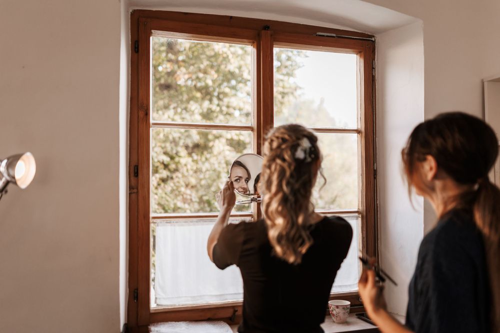 Braut betrachtet sich im Spiegel und prüft ihr Make-up, während hinter ihr die Visagistin steht und ebenfalls in den Spiegel blickt.
