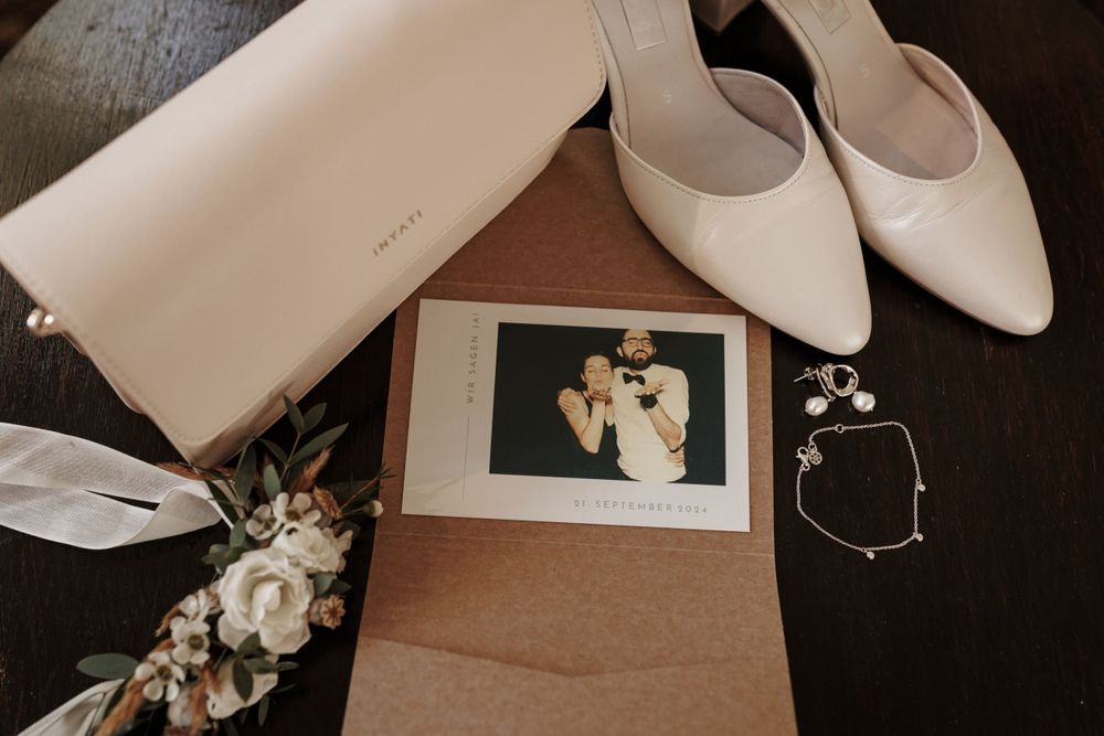 Hochzeitsdetails im Flatlay mit Brautschuhen mit weißen Absätzen, Clutch-Geldbörse, Vintage-Foto und zartem Blumenarrangement.