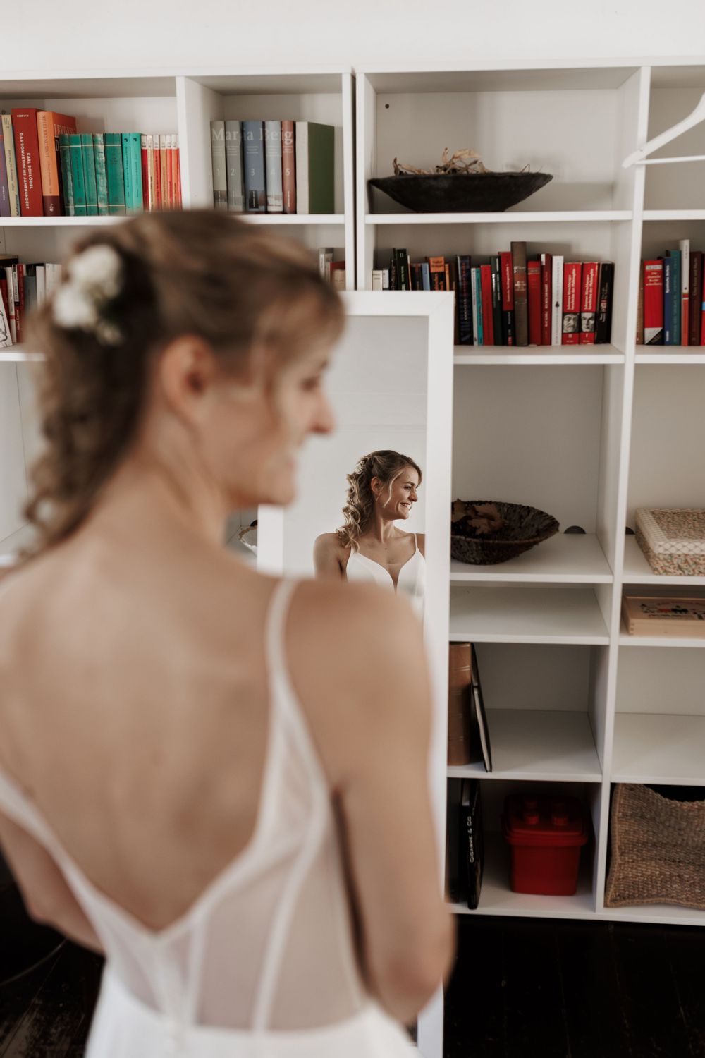 Eine Braut blickt in einen Spiegel in einem Raum mit weißen Bücherregalen voller bunter Bücher.