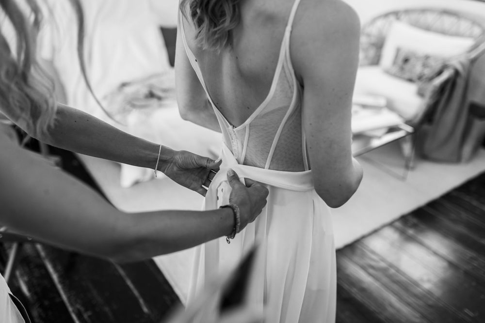 Schwarz-Weiß-Foto von jemandem, der von hinten dabei hilft, ein weißes Hochzeitskleid zu schnüren.