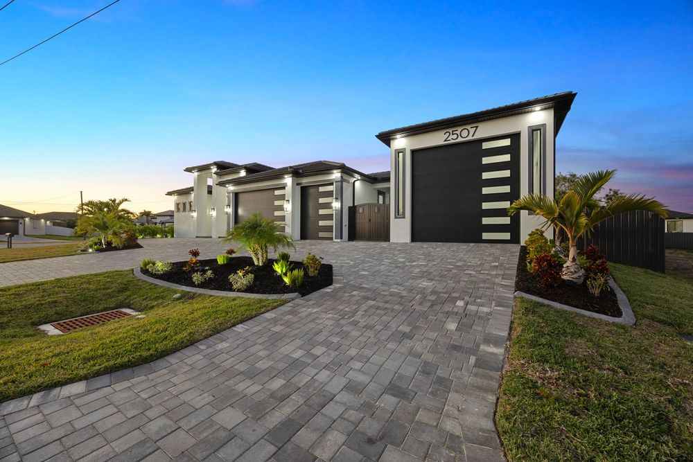 Modern two-story home with paver driveway, lush landscaping, and sleek garage door at dusk.