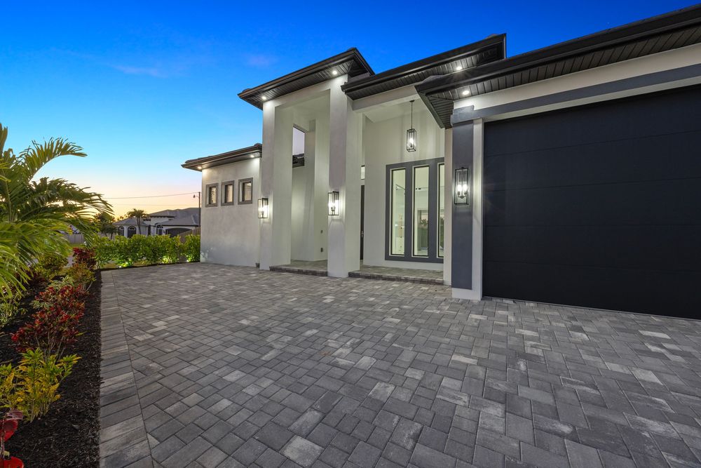 Modern luxury home exterior at dusk featuring paver driveway, dark garage door, and elegant glass entry doors.