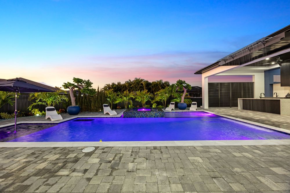 Luxury backyard pool with purple LED lighting, lounge chairs, lush landscaping, and outdoor kitchen at twilight.