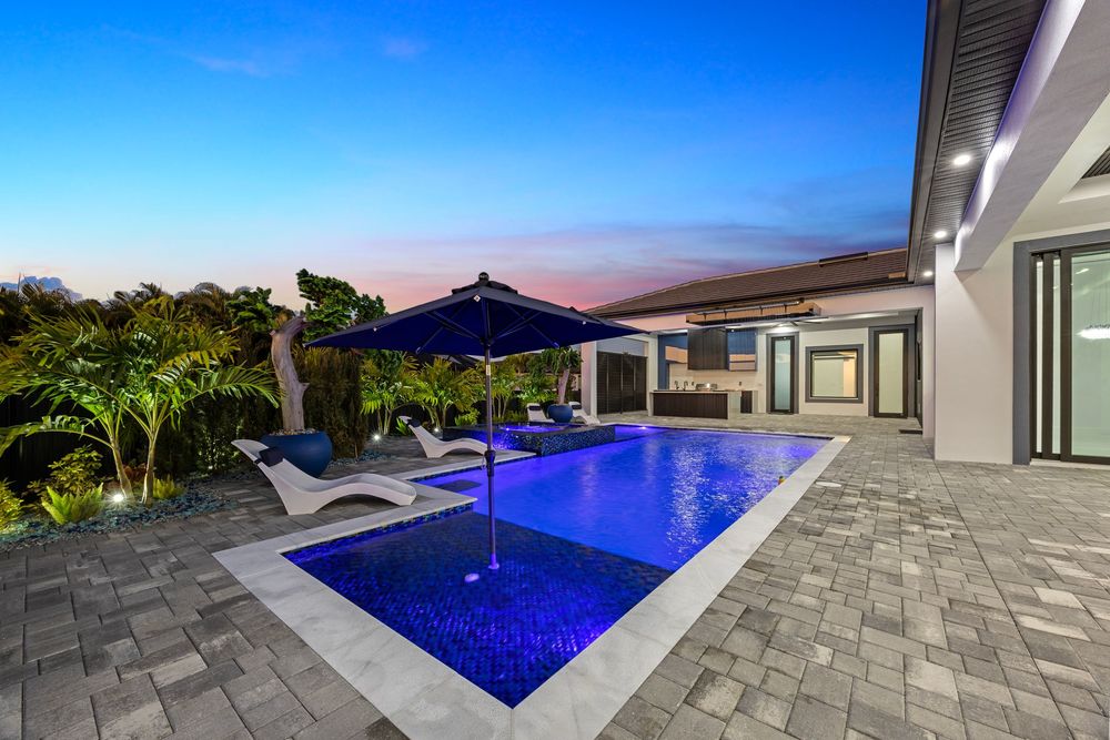 Luxury backyard pool with blue mosaic tiles, umbrella, lounge chairs, and lush landscaping at twilight.