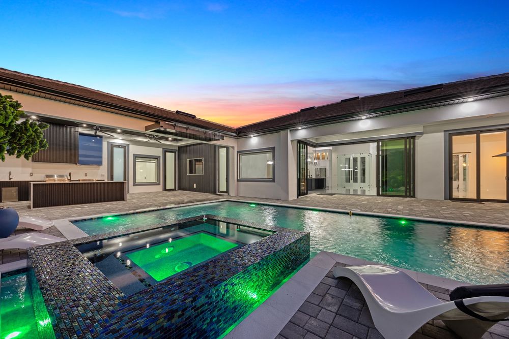 Elegant outdoor pool and spa with green LED lighting, lounge chairs, and modern home backdrop at dusk.