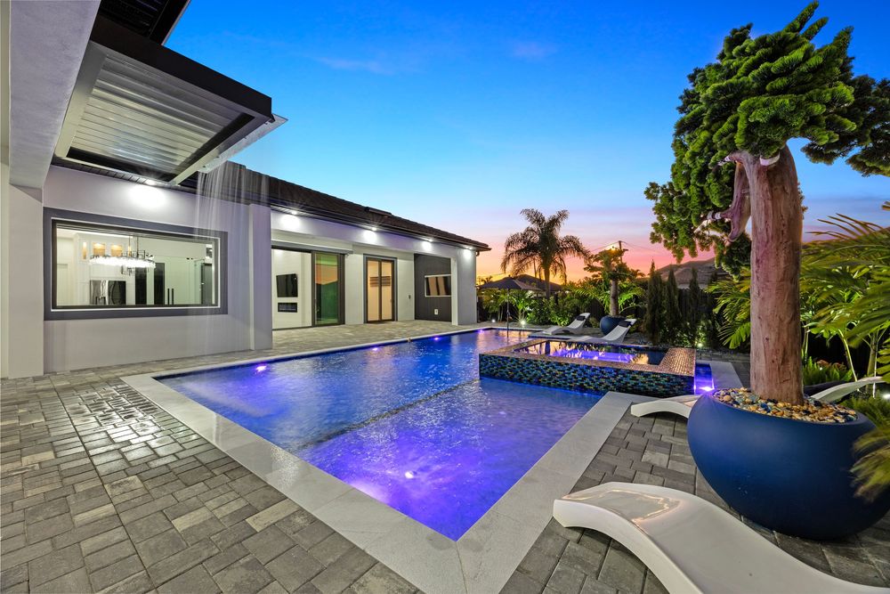 Luxurious backyard pool with blue LED lighting, paver patio, fire feature, and tropical landscaping at twilight.