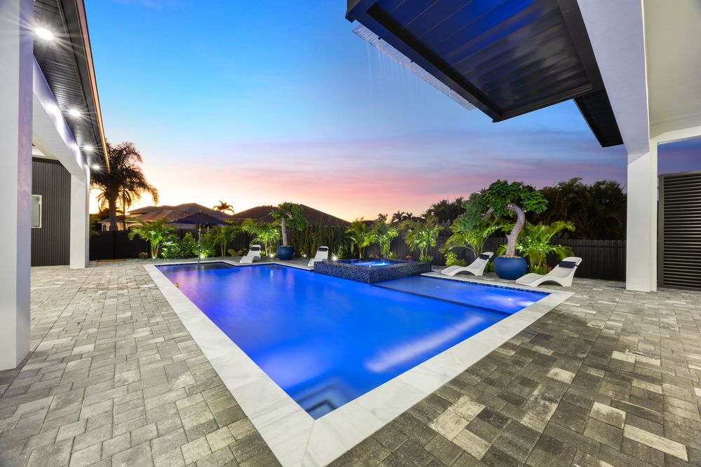 Luxury pool with glowing blue water, lounge chairs, and a stunning sunset sky in a modern backyard patio.