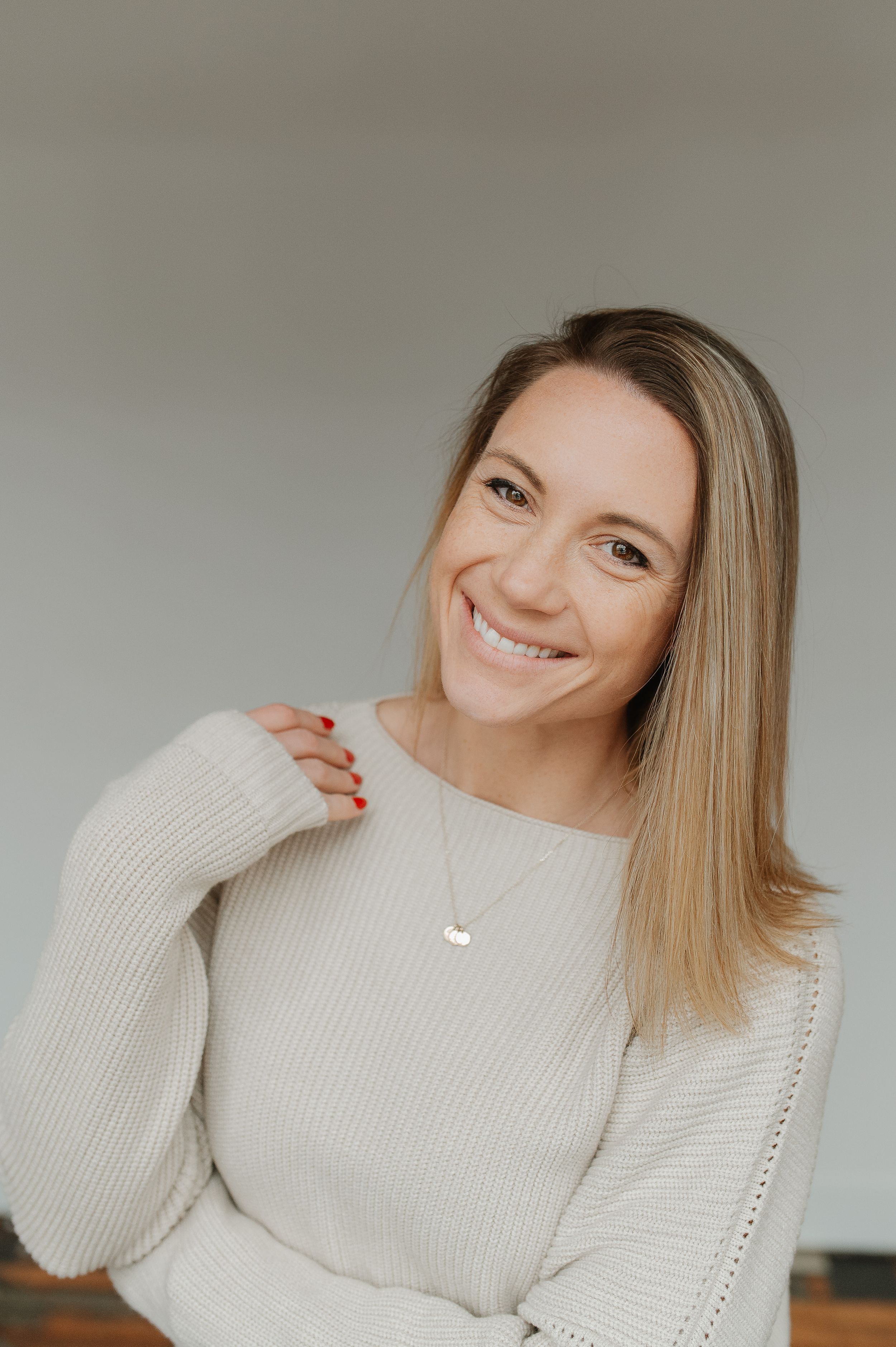 Portrait of someone wearing a cream-colored knit sweater with straight blonde hair and a delicate necklace.