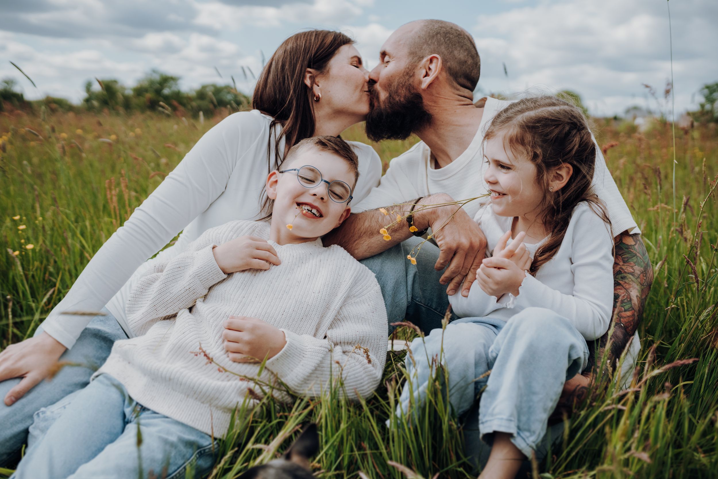Eine Familie sitzt glücklich auf einer Sommerwiese. Die Eltern küssen sich. Die Geschwister lachen.