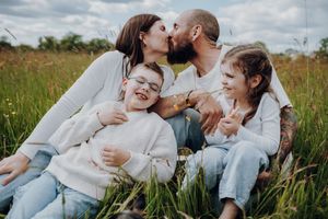 Eine Familie sitzt glücklich auf einer Sommerwiese. Die Eltern küssen sich. Die Geschwister lachen.