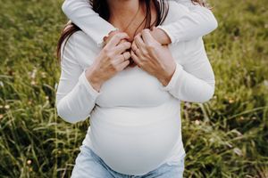 Die Tochter umarmt ihre Mama von hinten. Sie ist schwanger. Die Sonne scheint. Sie sind auf einer Sommerwiese.