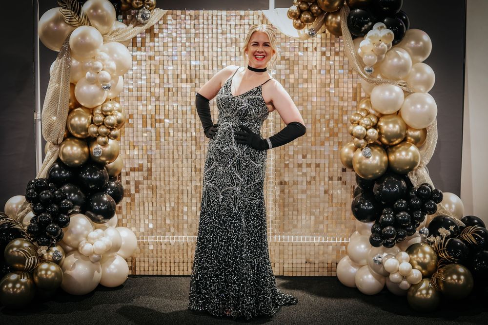 Formal event backdrop with gold and black balloon arch against sparkly wall.