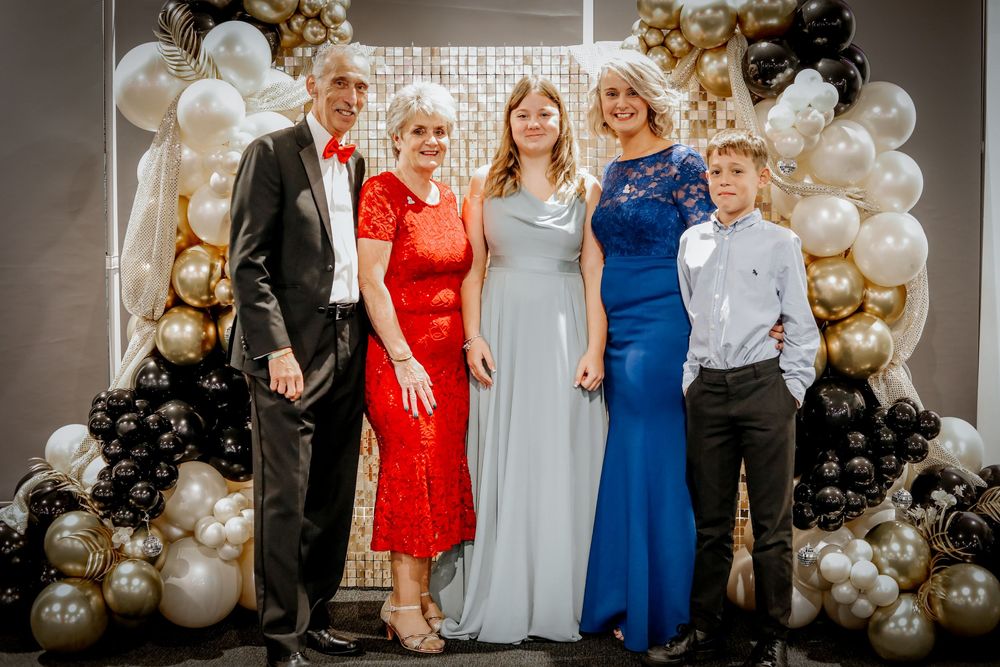 Formal group photo with balloon arch and sparkly backdrop at evening event.