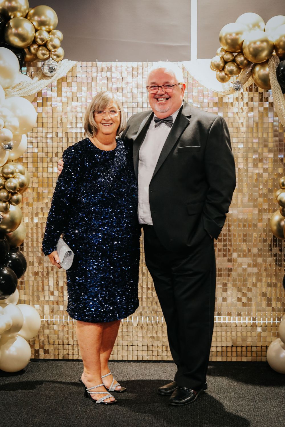 Formal couple posed against sparkly backdrop with gold and black balloon decorations.