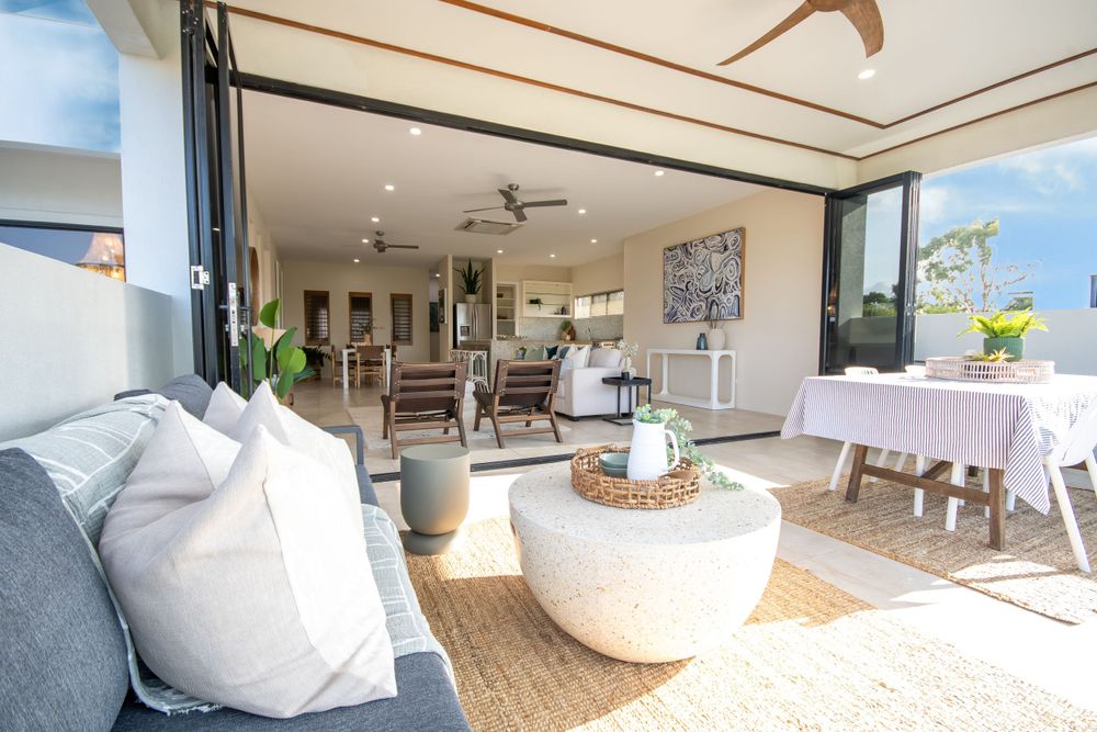 Coastal-inspired outdoor living space with woven furniture and white cushions opening to interior dining area.