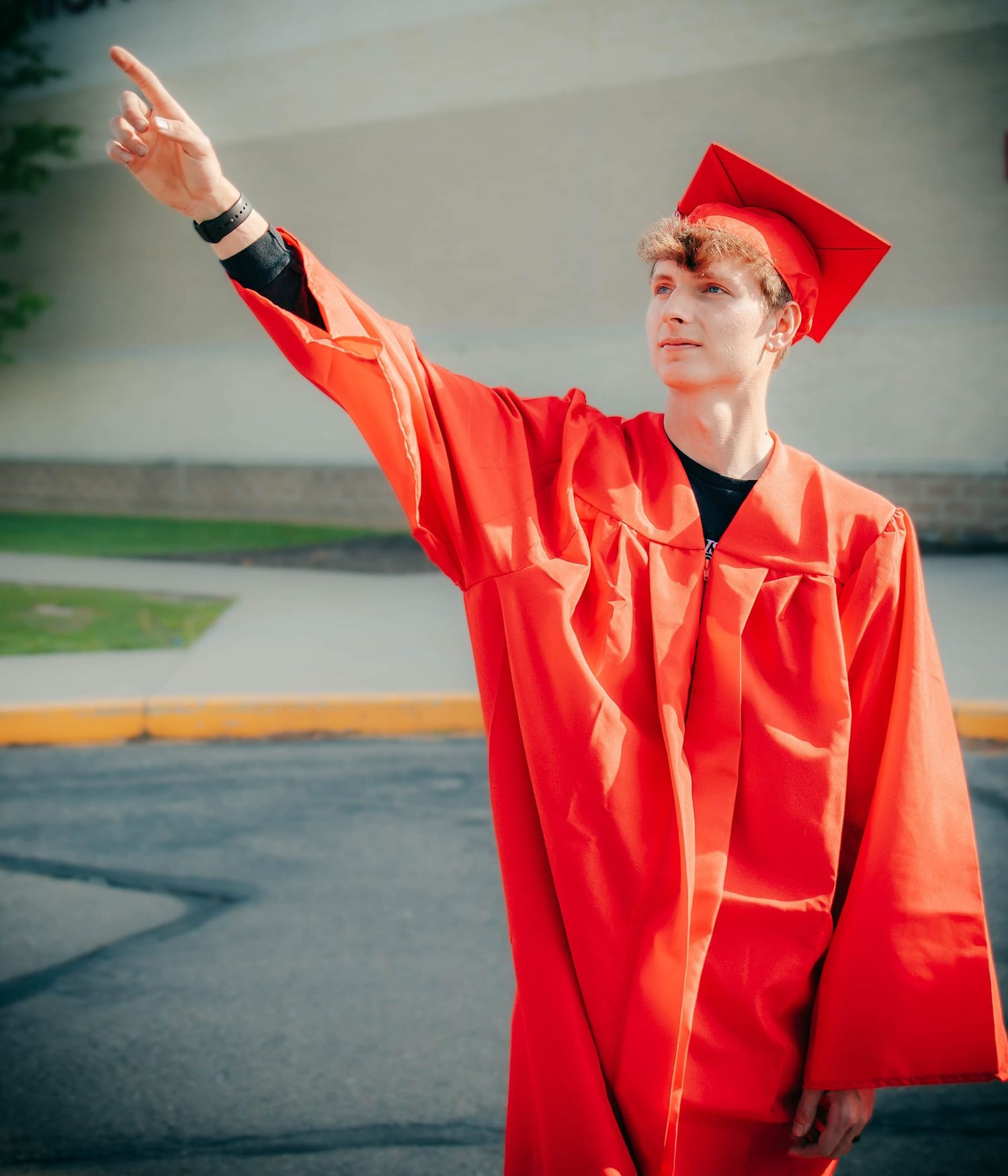 Graduate in bright red cap and gown pointing upward during outdoor ceremony.