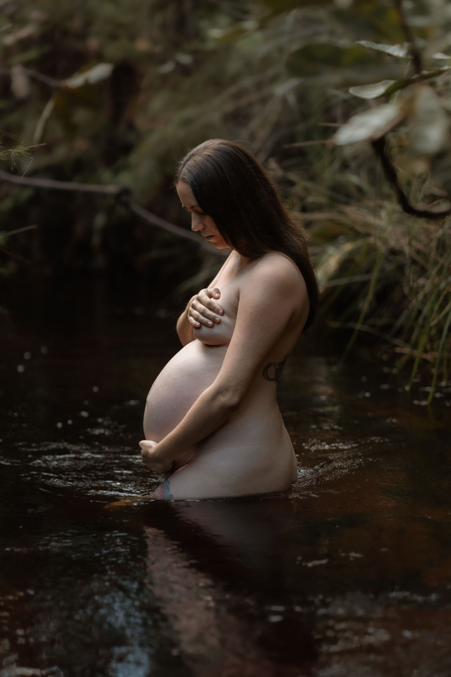 A pregnant woman sits serenely in dark water during a maternity photoshoot in a moody, natural setting.