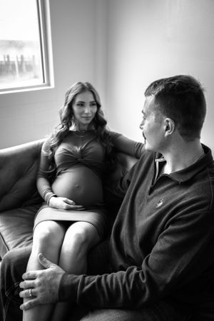 A black and white maternity photo with a couple sitting together on a couch in a softly lit room with a window. Maternity, Family, Studio