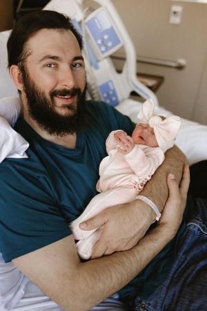 A bearded man in a navy blue shirt holds a sleeping newborn baby in a pink outfit in a hospital room. Fresh 48 Photography