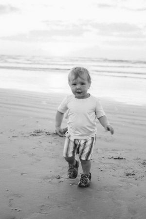 Young child wearing white t-shirt and striped shorts walks along a misty beach in black and white. Family photography