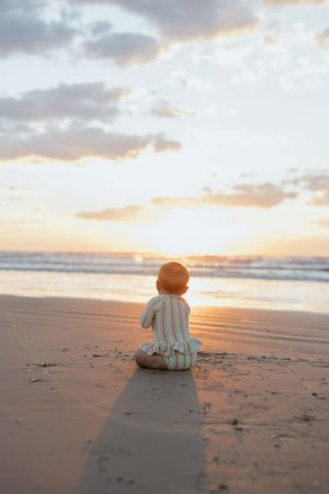 Silhouette of small child sitting alone on sandy beach watching beautiful orange sunset over ocean waves. Family Photography, Children