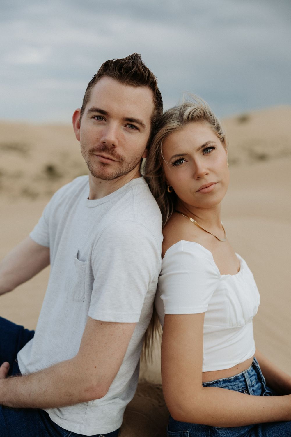 A casual couple poses back-to-back in white shirts against desert sand dunes.