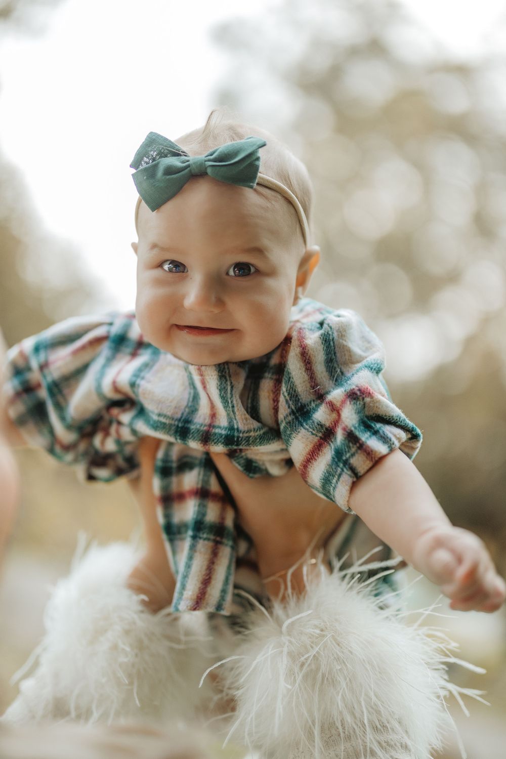Baby wearing plaid outfit and mint bow headband smiles at camera.
