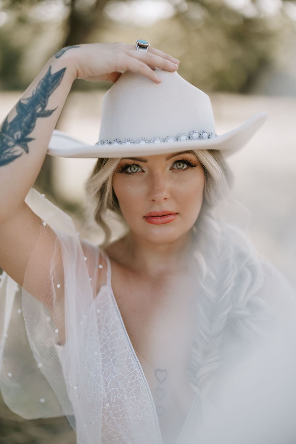 A dreamy portrait featuring a white cowboy hat, lace dress and visible arm tattoo in soft lighting.