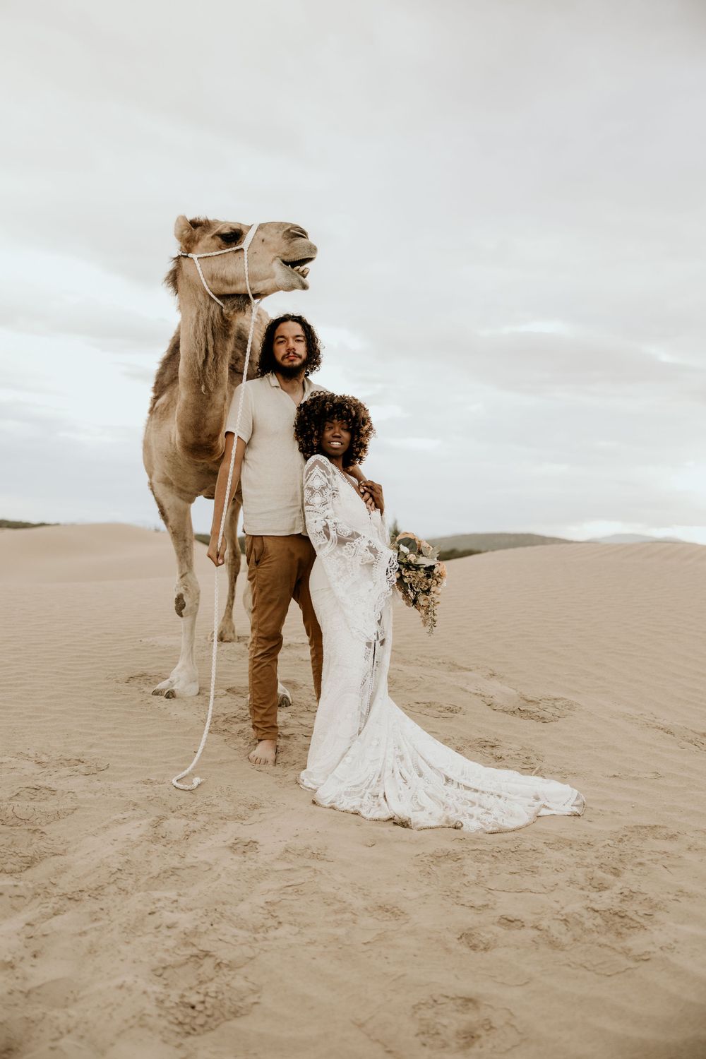 A desert wedding portrait featuring a couple and their camel against sandy dunes in sepia tones.
