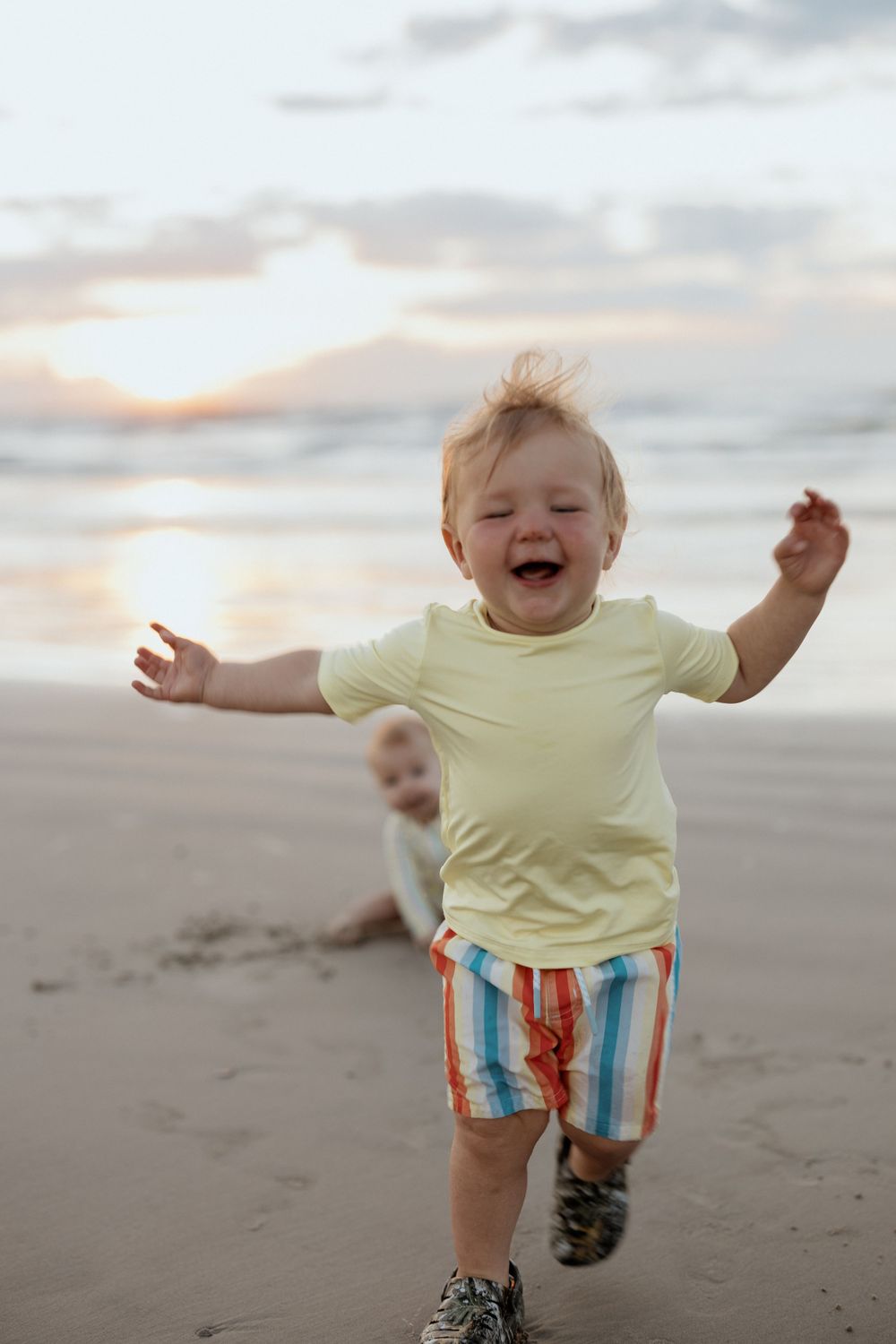 A joyful toddler runs and laughs on the beach during sunset wearing colorful striped shorts.