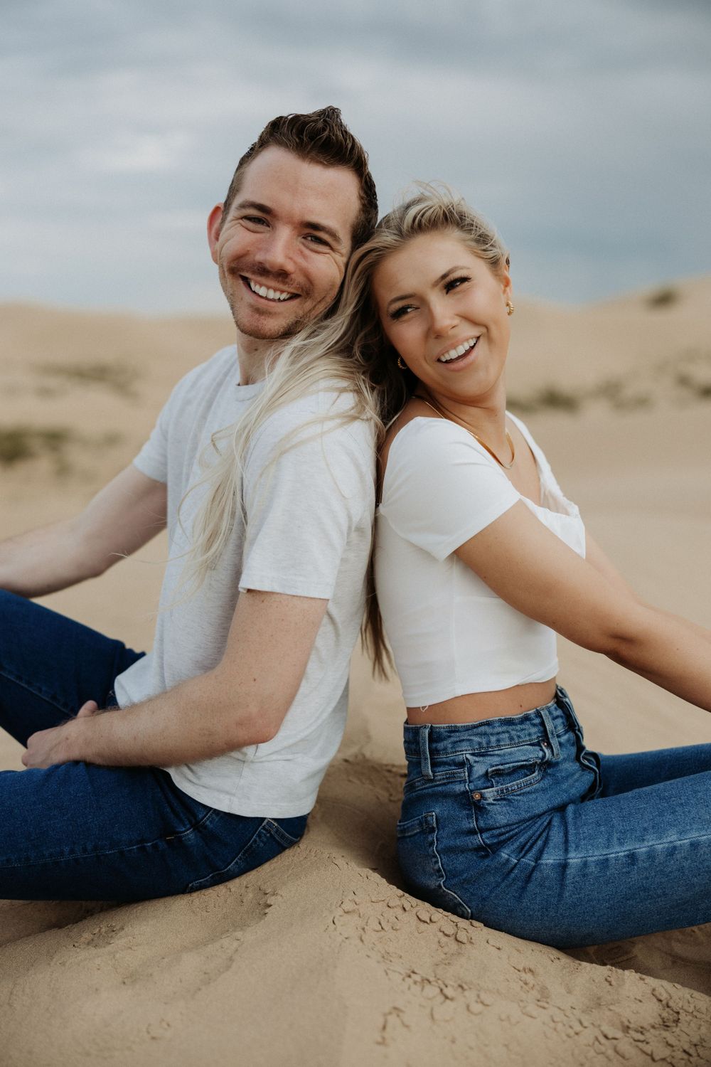 Candid couple sitting on sand dunes wearing casual white tops and jeans.