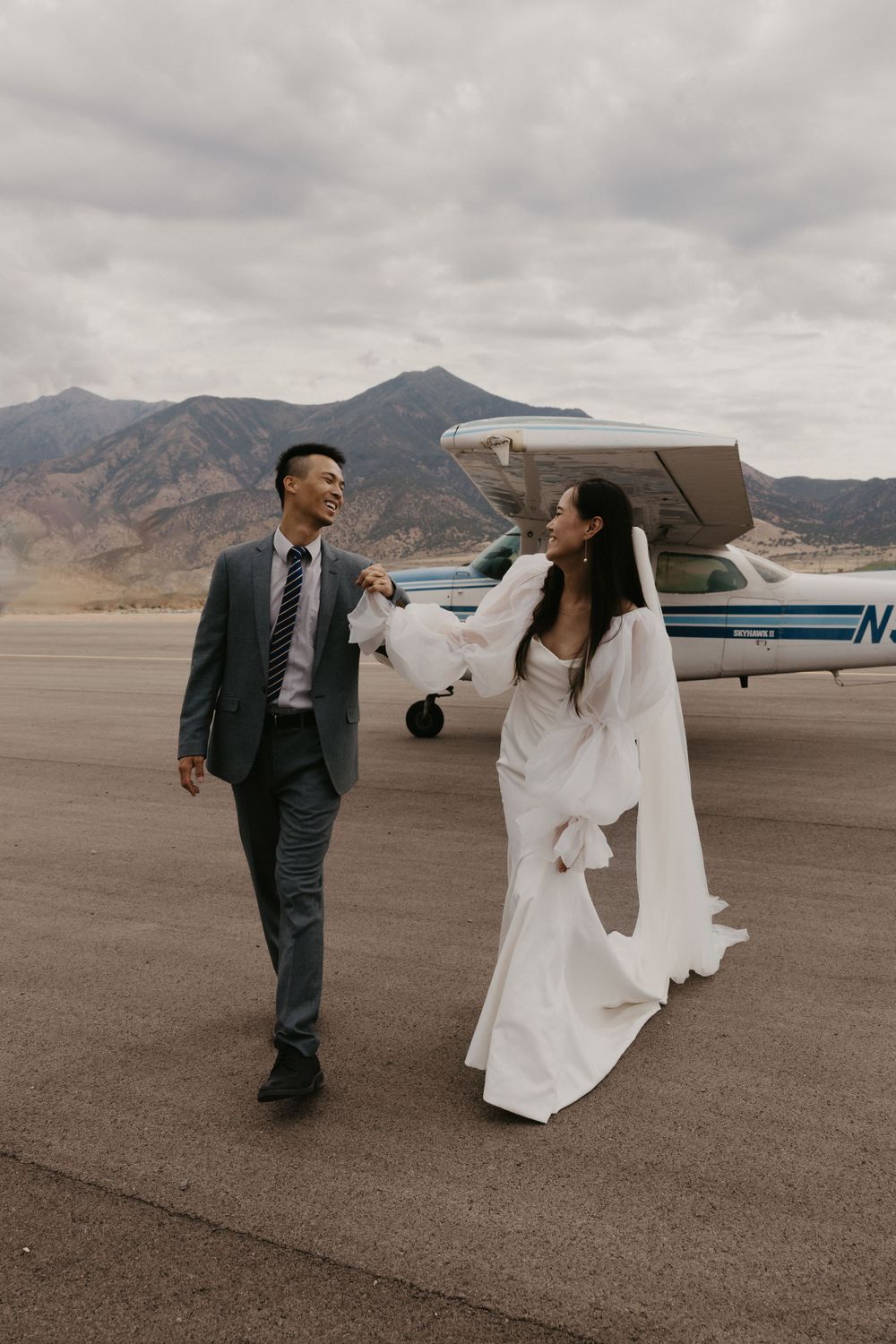 A wedding couple walks hand in hand on an airstrip with mountains and a small aircraft in the background.