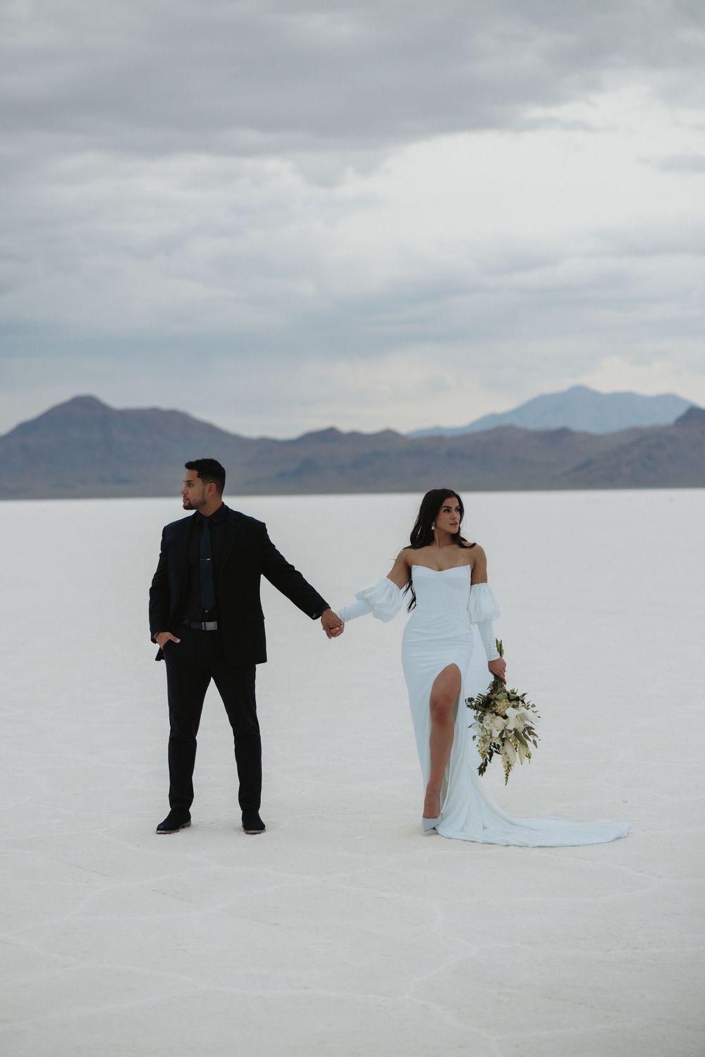 A stylish bride and groom pose dramatically on the stark white salt flats beneath cloudy mountain skies.