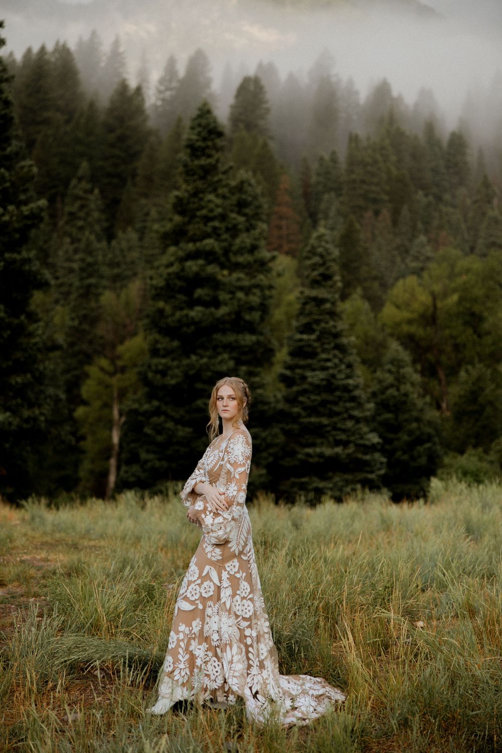 A ethereal bridal portrait in a misty forest meadow with towering evergreen trees in the background.