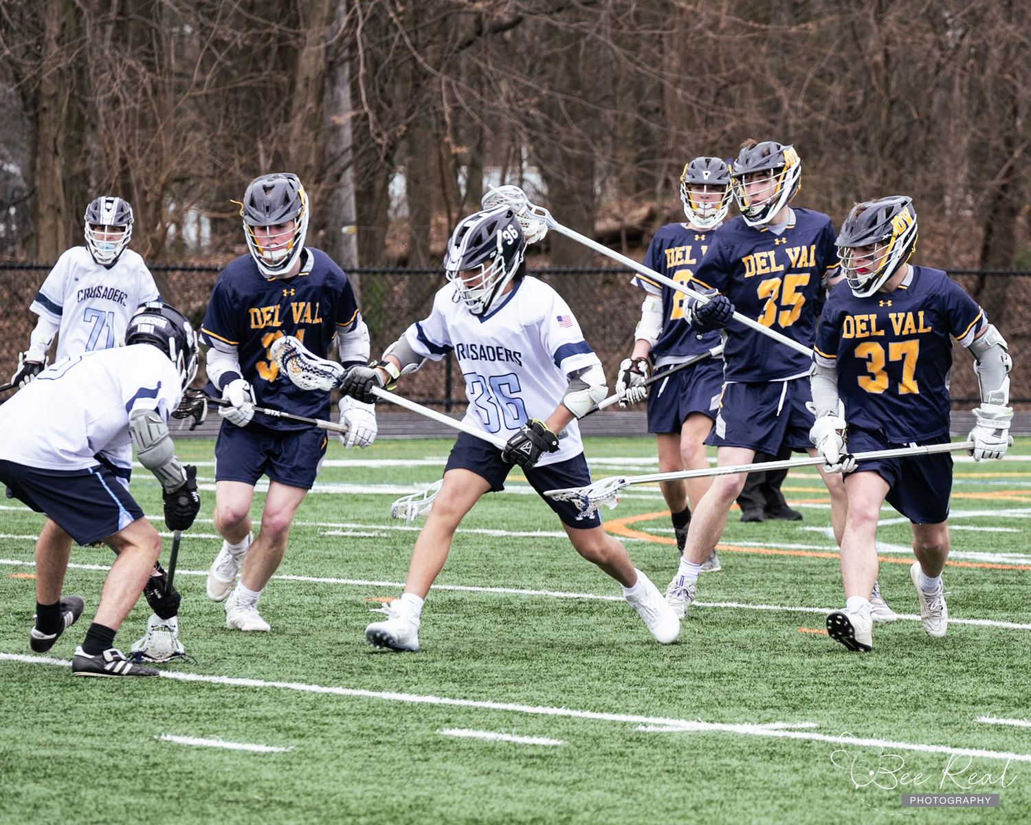 Lacrosse players in white and navy uniforms competing during an intense match on an artificial turf field.