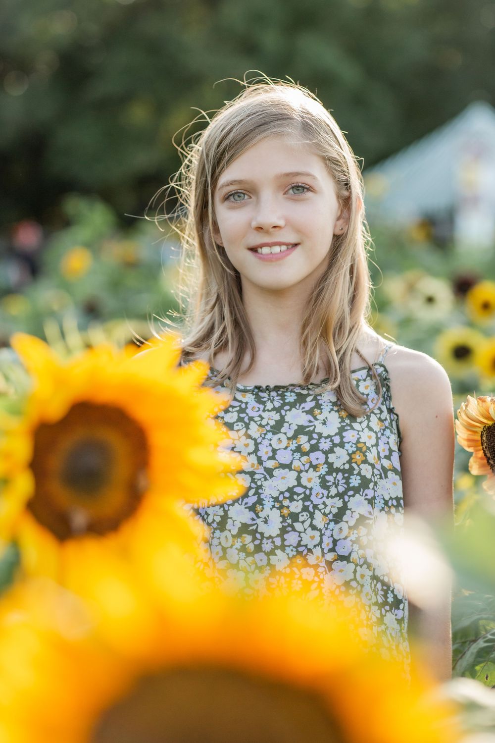 A smiling blonde girl in a floral dress stands among bright yellow sunflowers in a sunlit field.