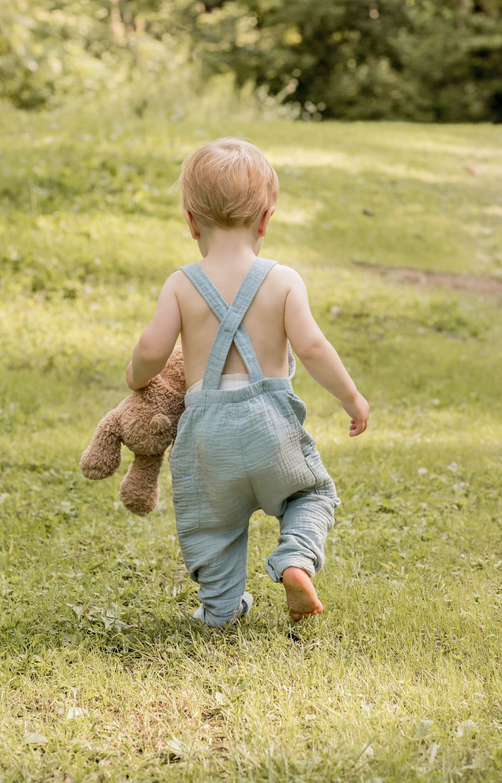 Toddler in blue linen overalls walking barefoot on grass carrying brown teddy bear.