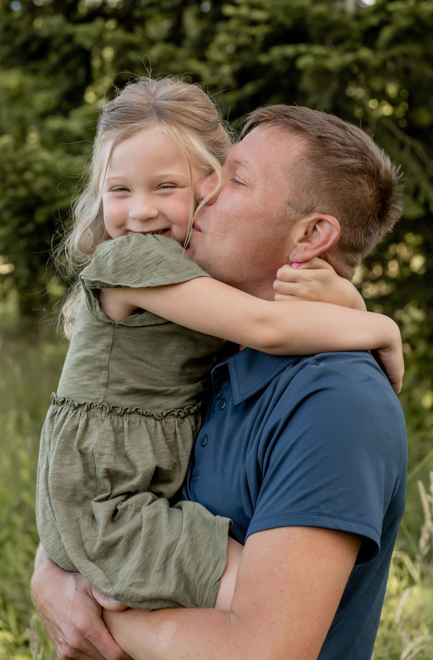 Father kissing cheek of smiling young daughter in green dress outdoors.