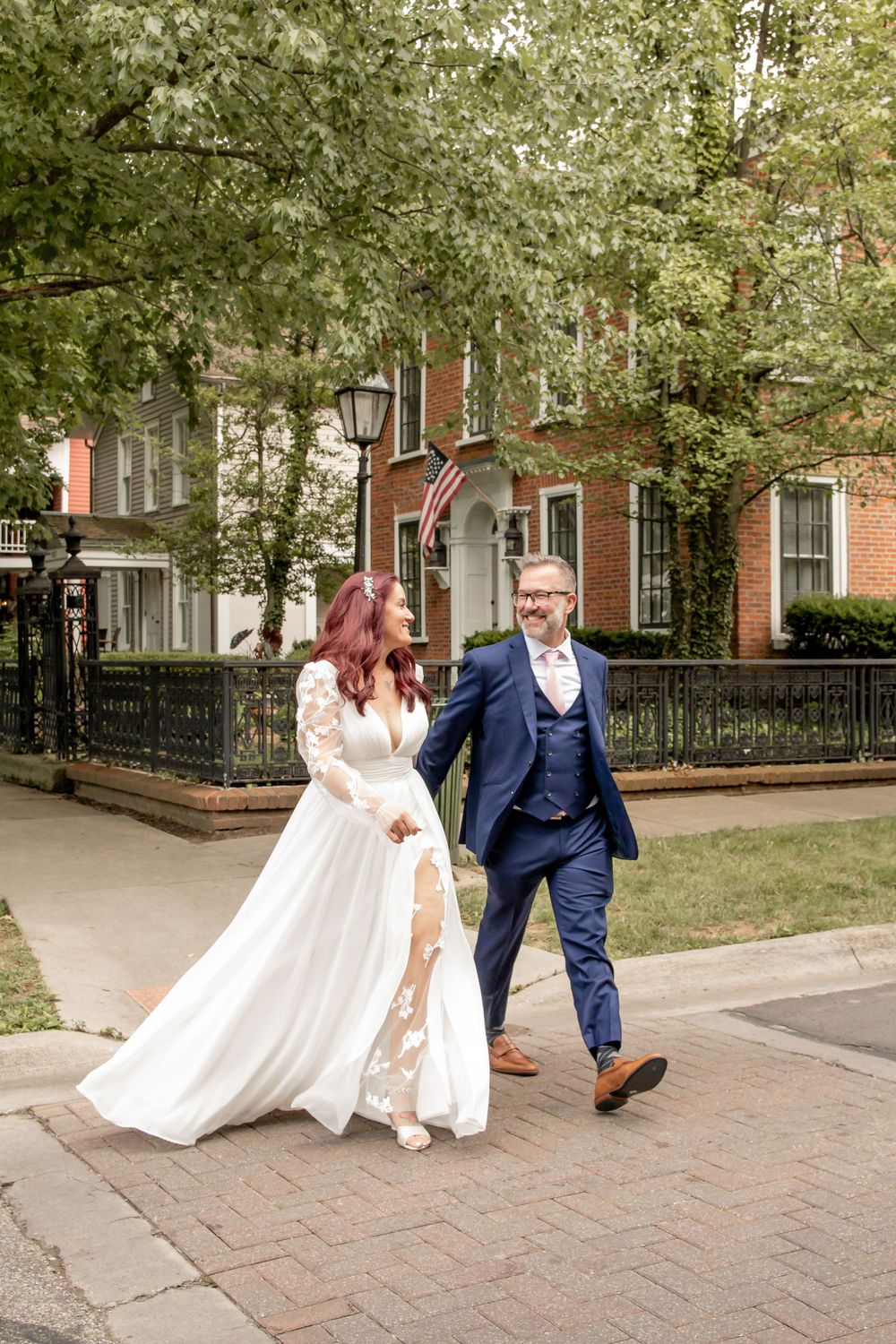 Joyful newlyweds walking hand in hand on brick sidewalk past elegant historic mansion.