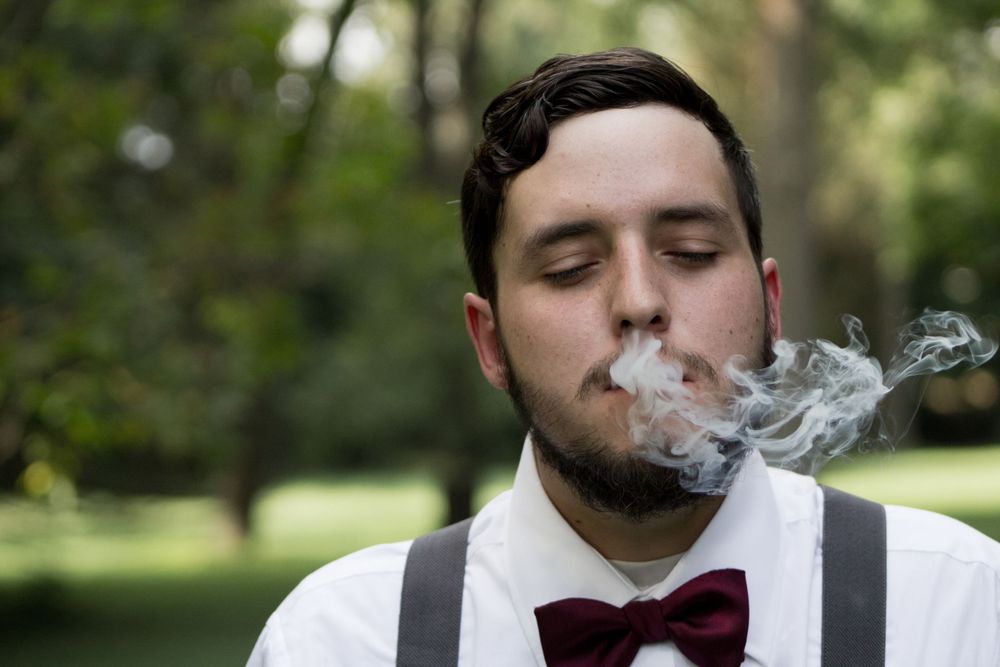 Young man in white shirt, suspenders, and burgundy bow tie exhaling smoke outdoors.
