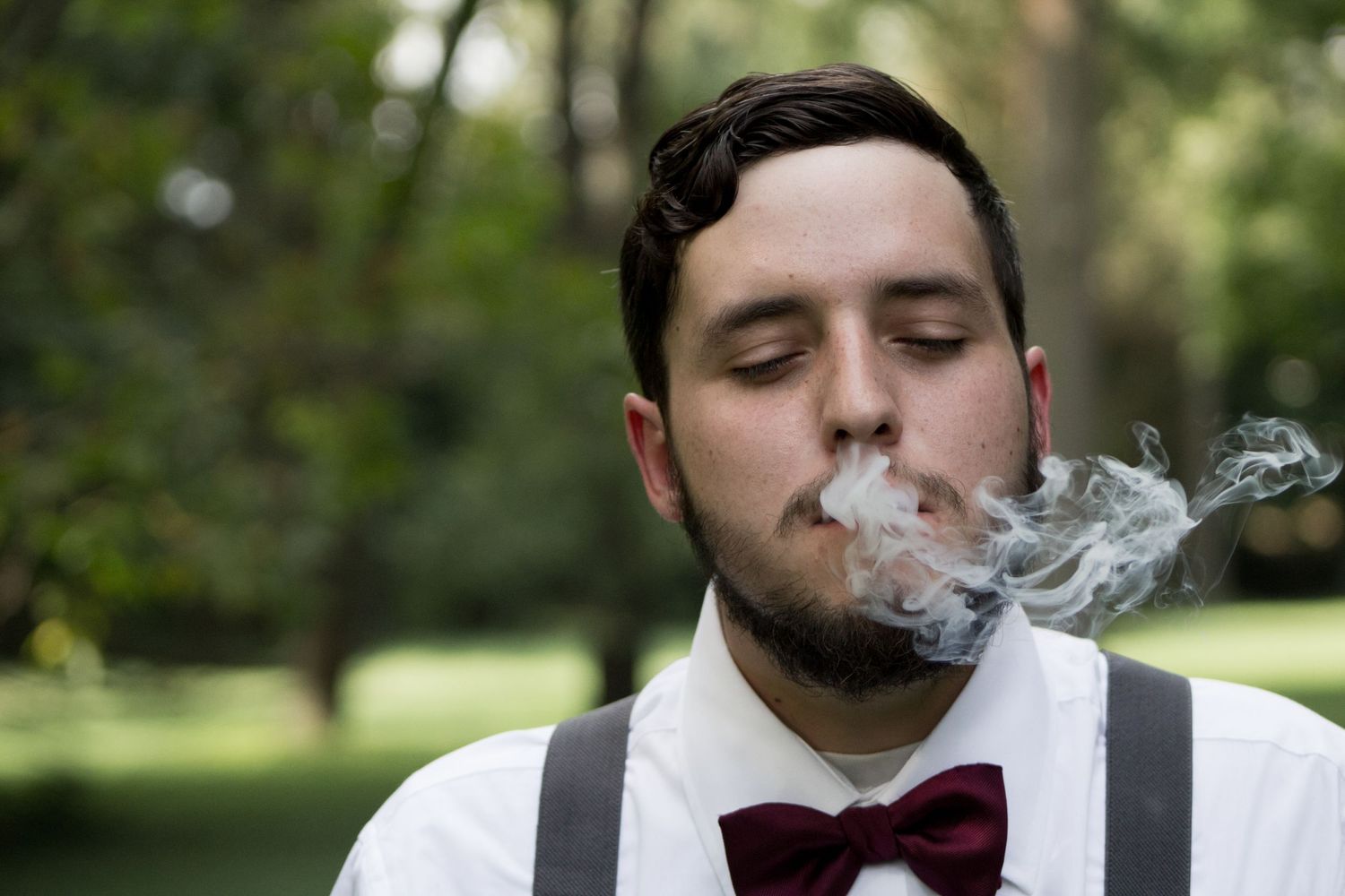 Young man in white shirt, suspenders, and burgundy bow tie exhaling smoke outdoors.
