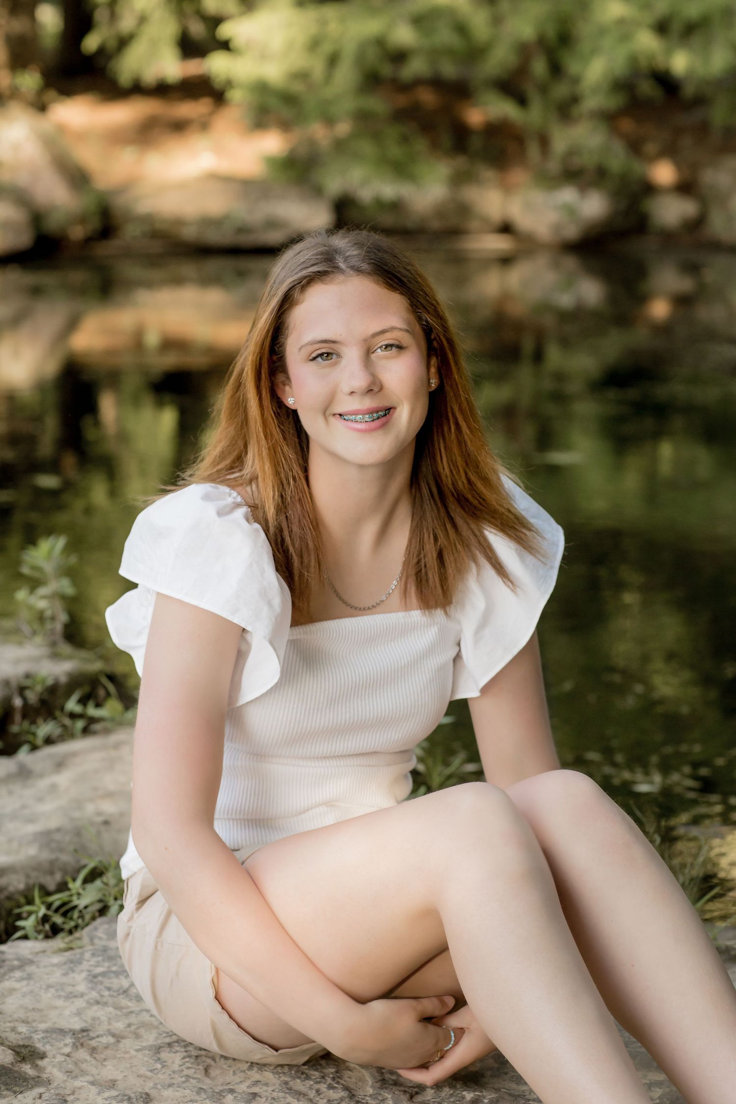 Smiling teenage girl with braces in white top sitting on rocks near a pond outdoors.