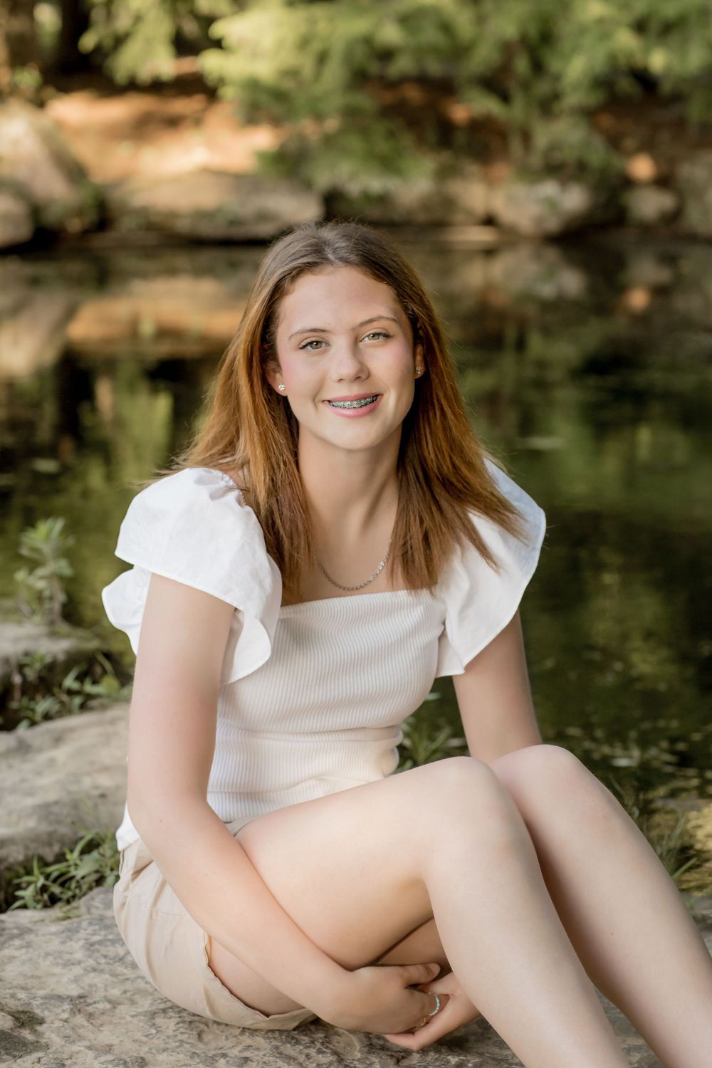 Smiling teenage girl with braces in white top sitting on rocks near a pond outdoors.