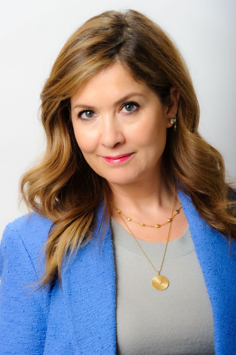 Professional headshot of woman in blue blazer and gray top with layered gold necklaces.