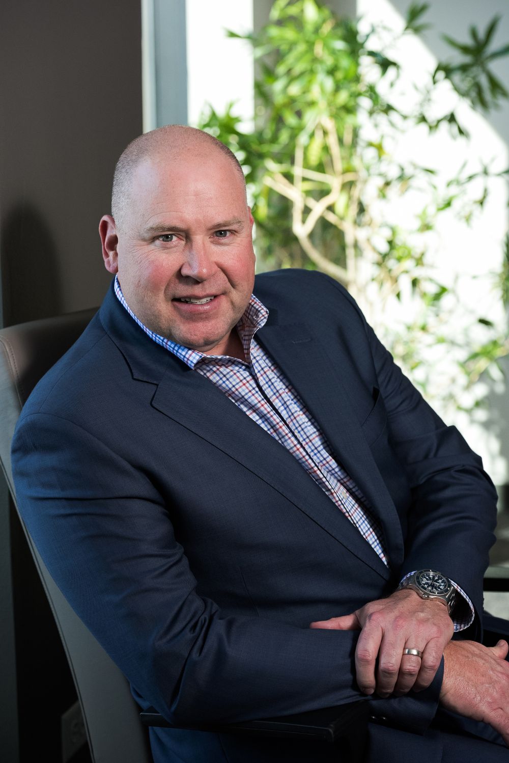 Business portrait of man in navy suit and checkered shirt seated by window with plants.